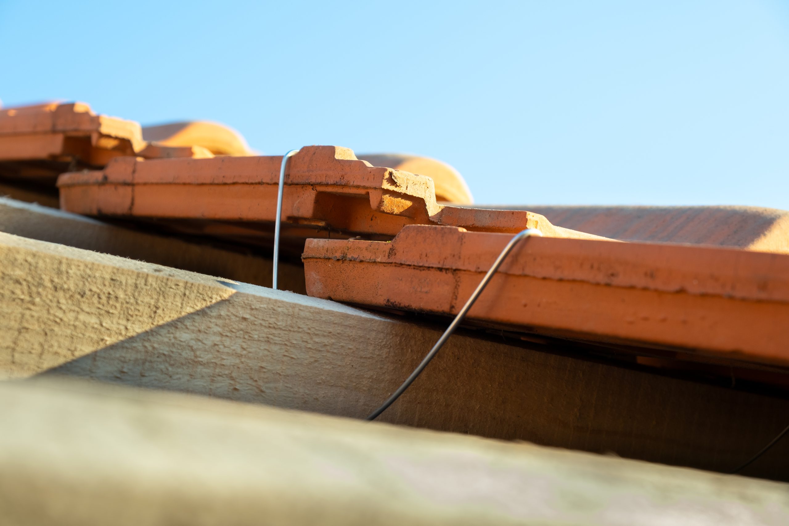 Closeup of metal montage anchor for installation of yellow ceramic roofing tiles mounted on wooden boards covering residential building roof under construction.